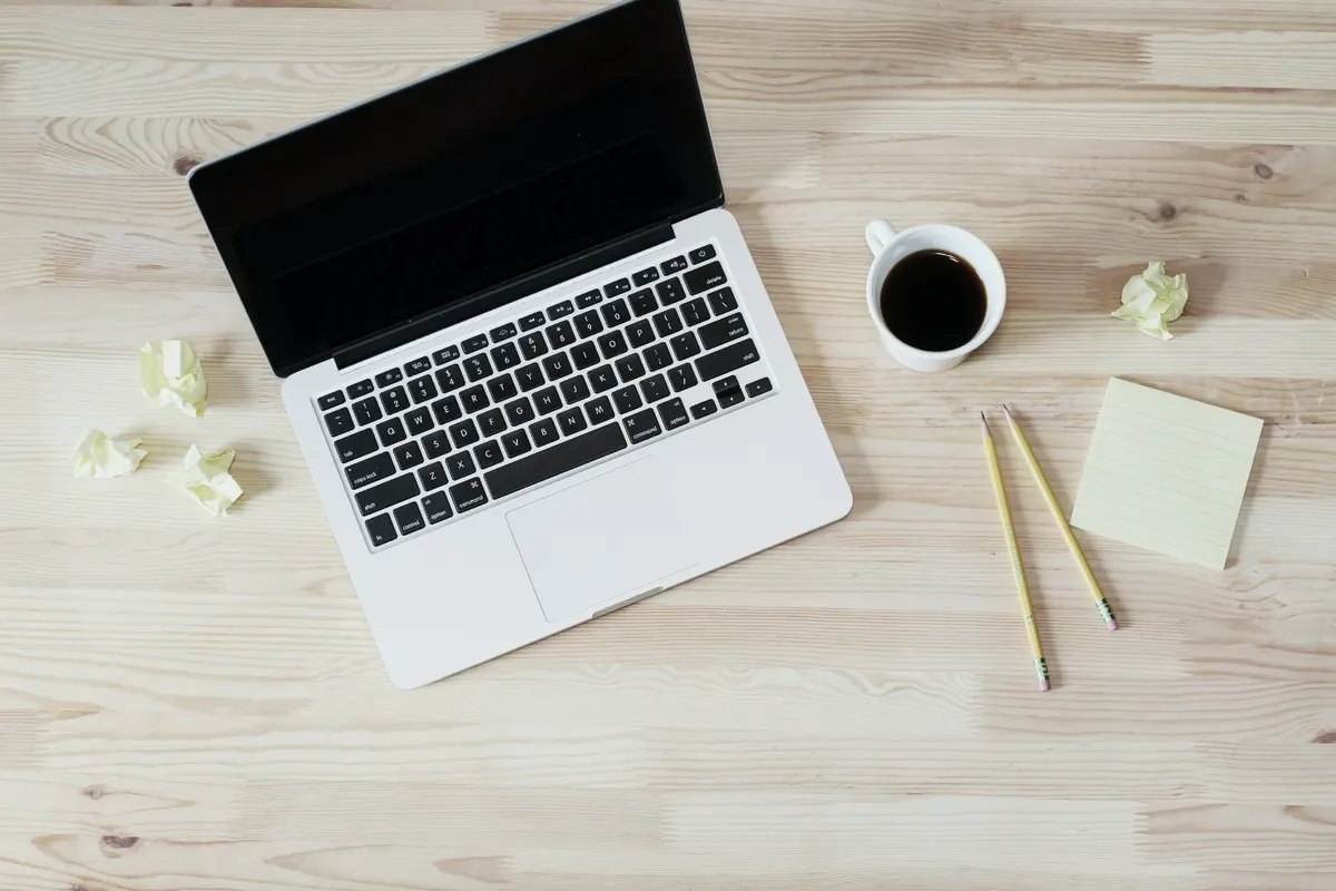 A laptop, a cup of coffee, pencils and a sticky notepad on a pale wooden desk