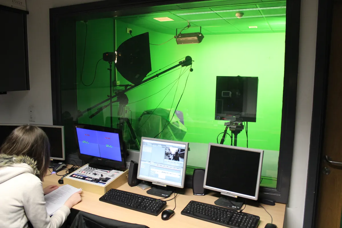 A small television studio seen through a control room window, with green screen, lights and monitors