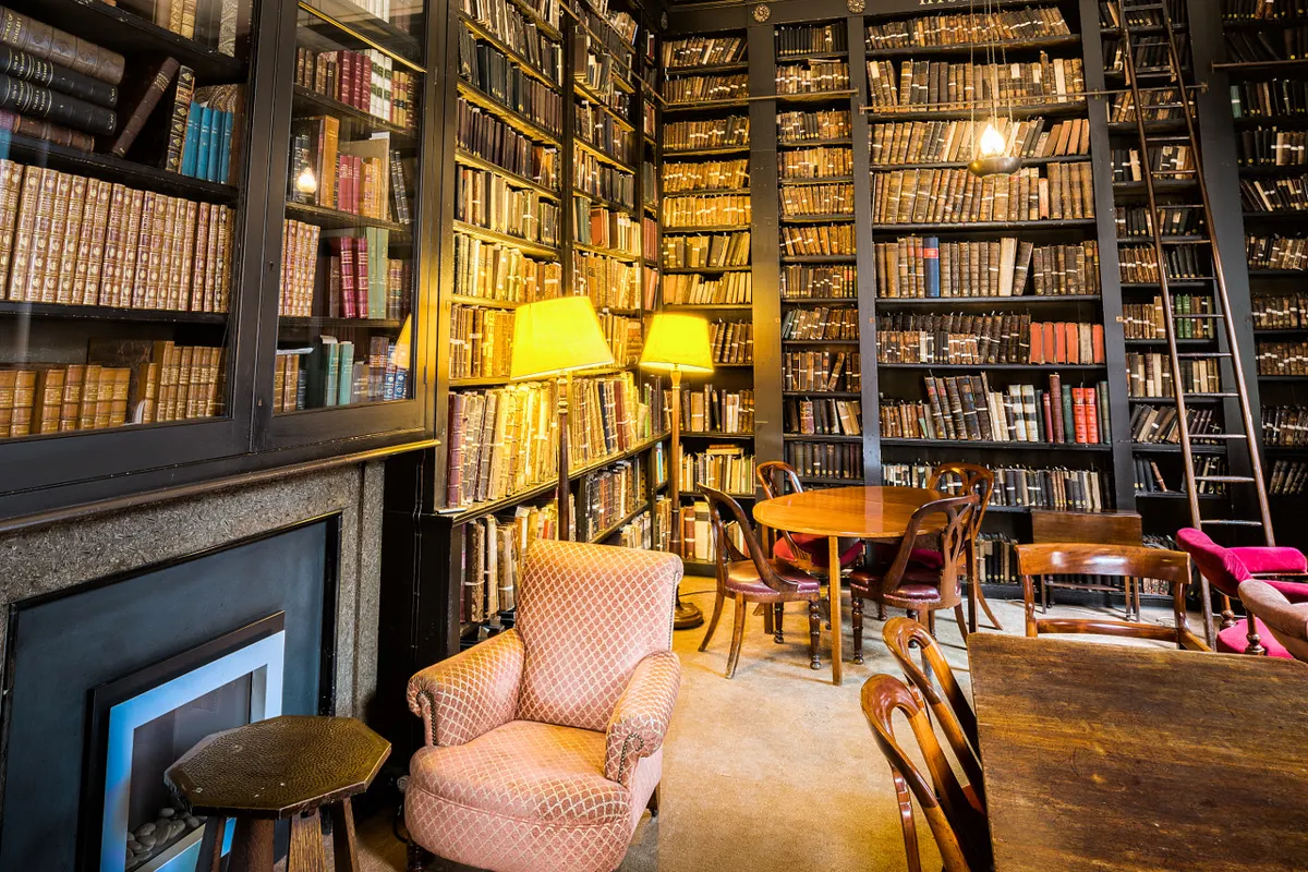 A quiet library reading room lined with shelves of old books, armchairs and a wooden table