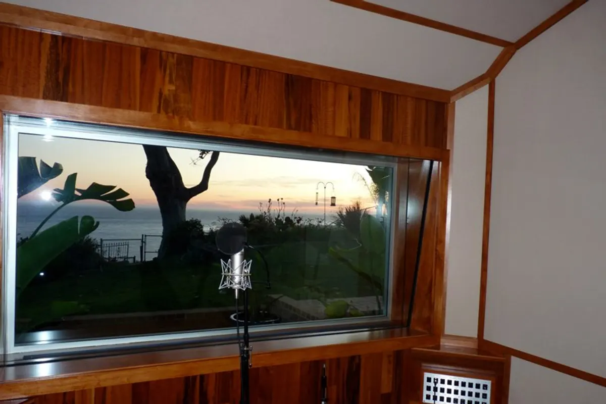 A condenser microphone on a stand inside a wood-panelled vocal booth with a window onto a garden at dusk
