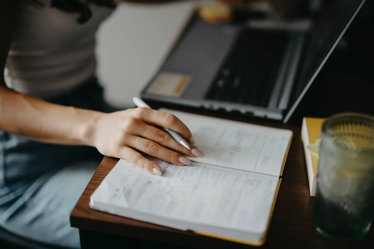 A person's hand resting on an open handwritten notebook beside a laptop on a desk
