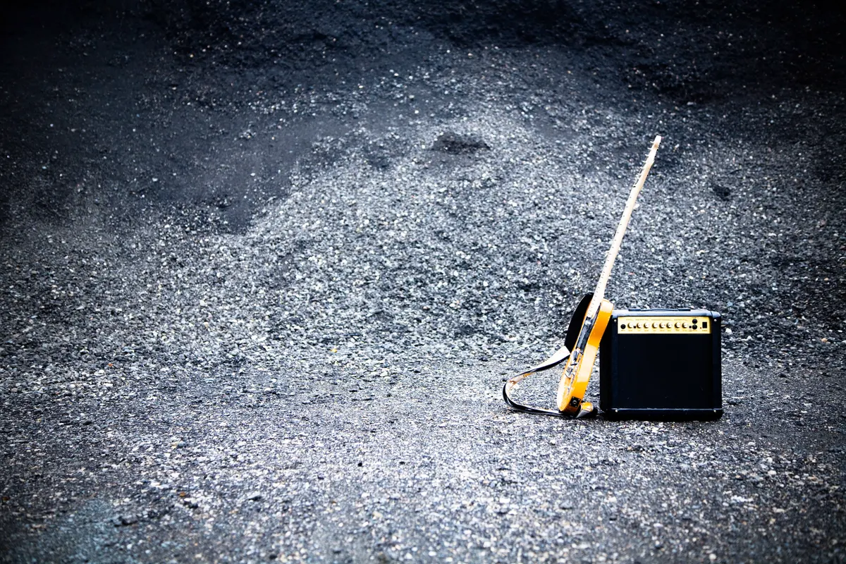A yellow electric guitar leaning against a small black practice amplifier on gravel