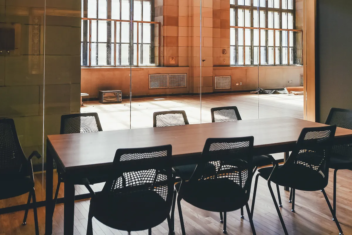 An empty conference room with a long wooden table and black chairs beside tall windows