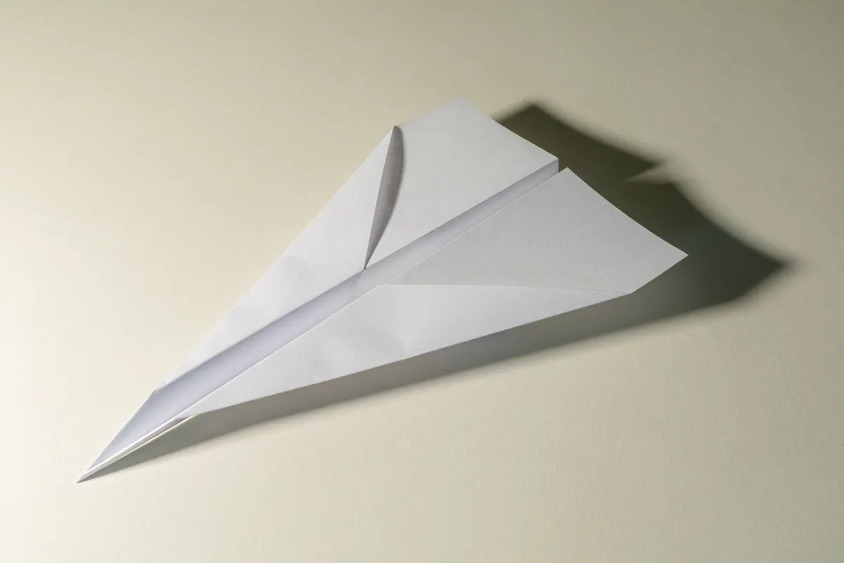 A white folded paper airplane on a plain surface with a soft shadow