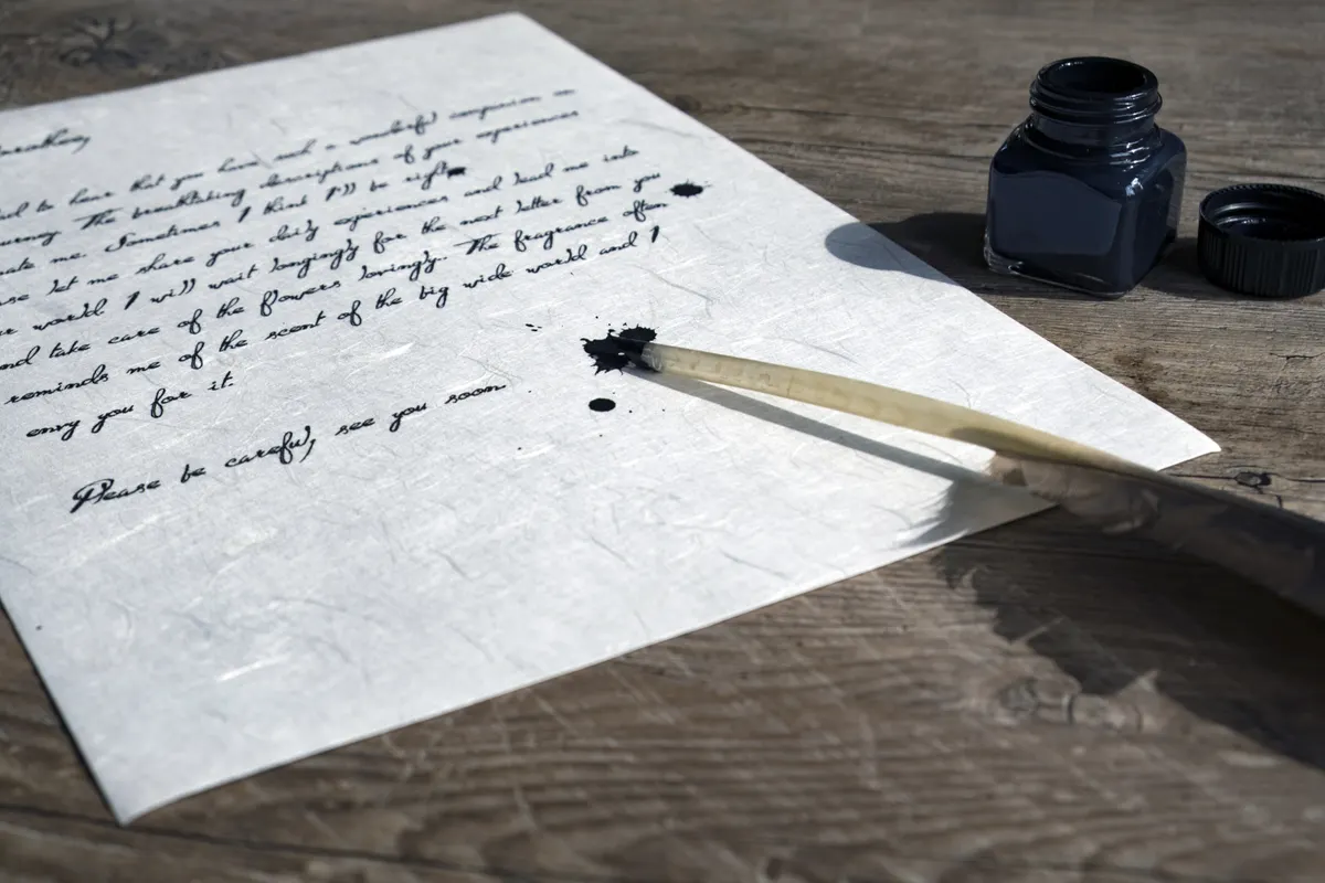 A handwritten letter on pale paper beside an ink bottle and a feather dipping pen