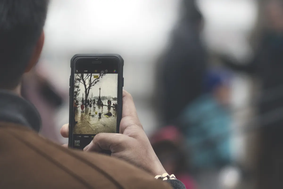 A hand holds a phone upright, filming a street scene shown on its screen