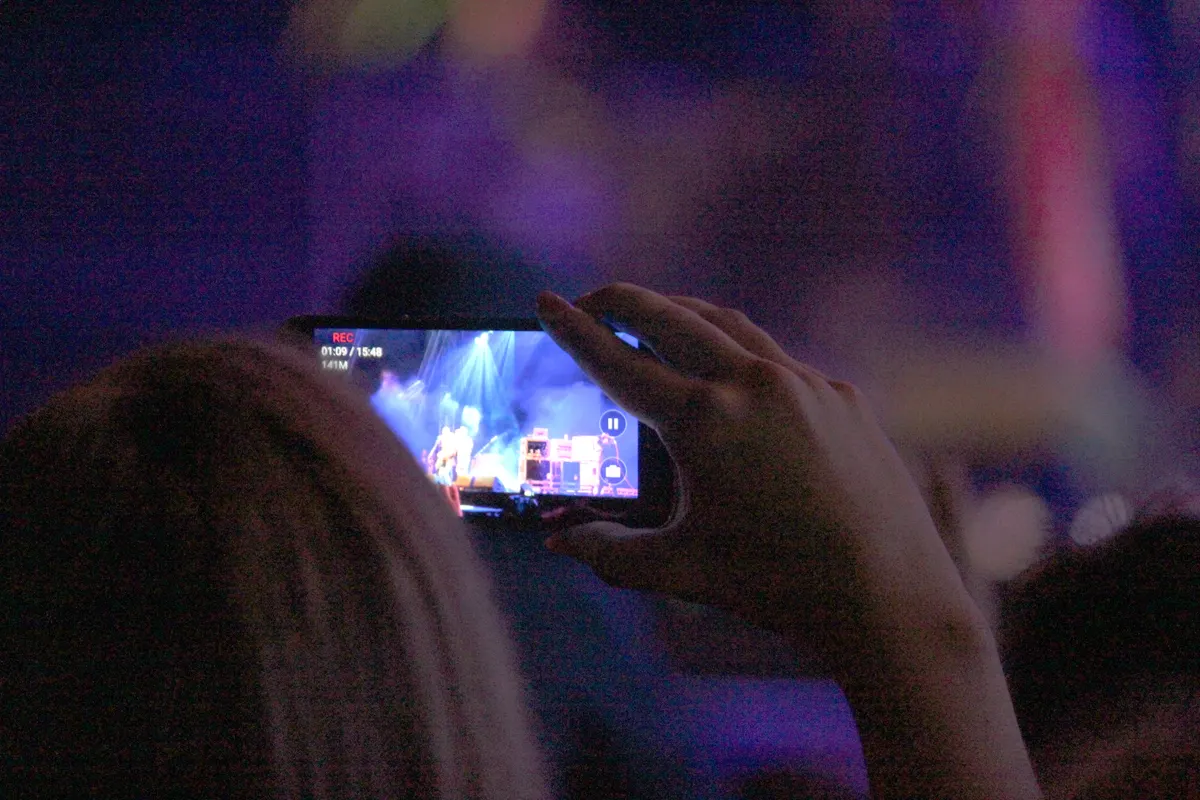 A hand holds a phone recording a lit stage from the dark of a crowd